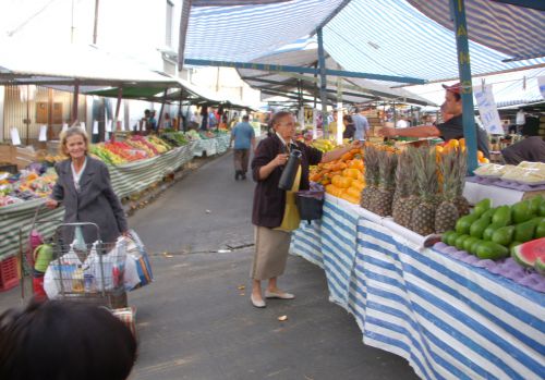 フェイラの店主にコーヒーを渡す女性と、保温ポットの入ったカゴを立てて待っている女性