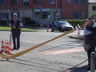 交通量の多い道路の脇で、民族衣装に身を包んだオジサン２人がアルペンホルン演奏中