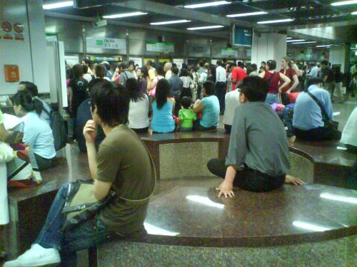 ラッシュ時の地下鉄のホーム。数台電車を見送る人々がベンチに座っている。