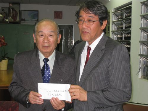 ブラジル熊本県文化交流協会の田呂丸会長（左）に義援金を手渡す梅田ブラジル大使